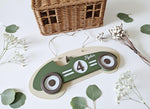 May include: A wooden race car-shaped sign in olive green and beige, with the name "Ayden" and the number "4" displayed. The wheels are brown and white. The sign is suspended by a string. Decorative elements include sprigs of white flowers and green leaves.