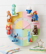 May include: A pastel-coloured children's wall clock with cartoon character figurines. The clock face is divided into pastel segments. The clock hands are silver, and the numbers are gold. The clock's diameter is approximately 30 cm.